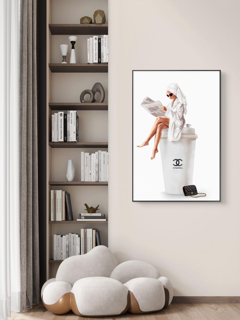 May include: A framed art print depicts a woman in a white robe and towel, reading a newspaper while seated on a large white coffee cup. The cup features the Chanel logo and a small black purse rests nearby. The artwork is displayed in a modern interior.