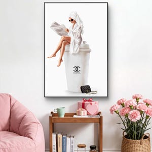 May include: A framed art print depicts a person in a white robe and towel reading a newspaper while seated on a large white coffee cup with the Chanel logo. A black purse and gold chain are at the base of the cup.