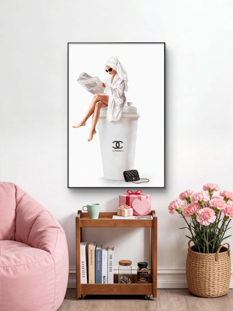 May include: A framed art print depicts a person in a white robe and towel reading a newspaper while seated on a large white coffee cup with the Chanel logo. A black purse and gold chain are at the base of the cup.