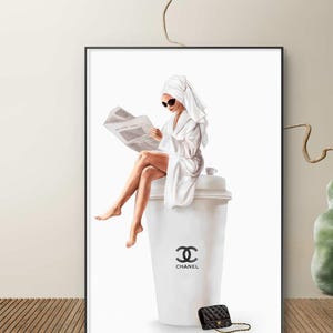 May include: A framed art print depicts a person in a white robe and sunglasses, reading a newspaper while seated on a large white coffee cup. A black purse with a gold chain sits below the cup. The cup features the Chanel logo.