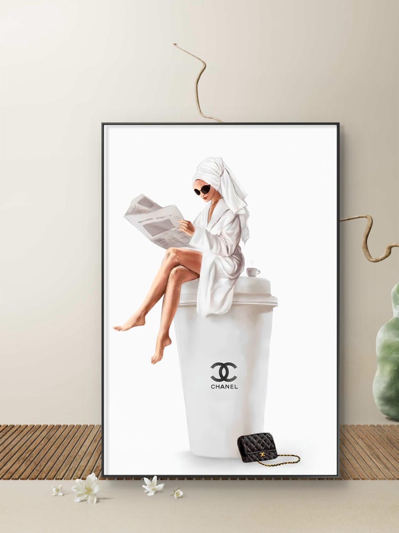 May include: A framed art print depicts a person in a white robe and sunglasses, reading a newspaper while seated on a large white coffee cup. A black purse with a gold chain sits below the cup. The cup features the Chanel logo.
