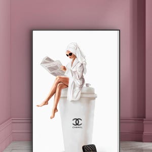 May include: A framed art print depicts a person in a white robe and towel reading a newspaper while seated on a large white coffee cup. The cup features the Chanel logo and text. A black quilted purse with a gold chain is at the base.