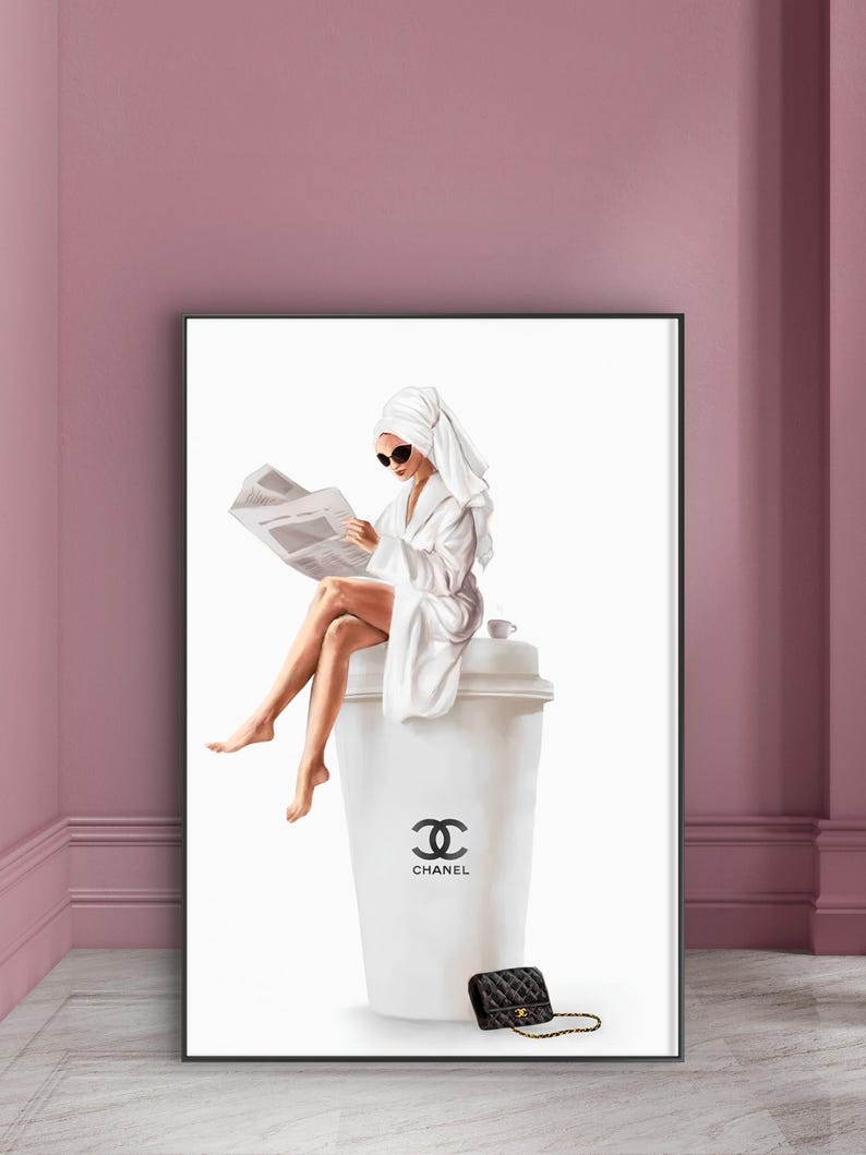 May include: A framed art print depicts a person in a white robe and towel reading a newspaper while seated on a large white coffee cup. The cup features the Chanel logo and text. A black quilted purse with a gold chain is at the base.