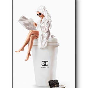 May include: A framed print depicts a person in a white robe and towel reading a newspaper while seated on a large white coffee cup with the Chanel logo. A small black quilted purse with a gold chain sits nearby.