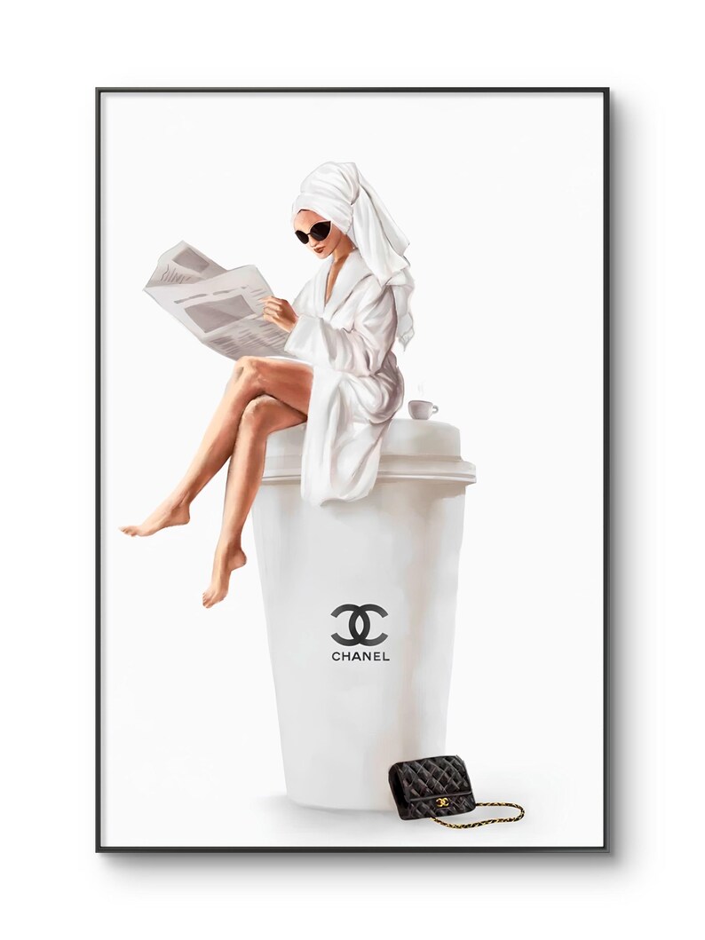 May include: A framed print depicts a person in a white robe and towel reading a newspaper while seated on a large white coffee cup with the Chanel logo. A small black quilted purse with a gold chain sits nearby.