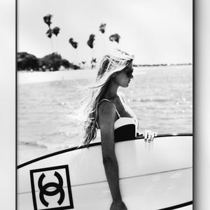 May include: Black and white framed print featuring a woman in a swimsuit holding a surfboard. The surfboard has a black logo on it. The background shows a beach scene with palm trees and the ocean.
