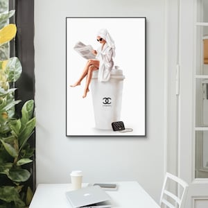 May include: A framed art print featuring a woman in a white robe and towel, reading a newspaper while seated on a large coffee cup. The cup has a black logo and the word "CHANEL". A small black purse with a gold chain is next to the cup.