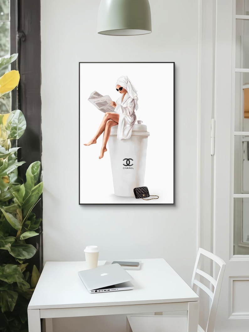 May include: A framed art print featuring a woman in a white robe and towel, reading a newspaper while seated on a large coffee cup. The cup has a black logo and the word "CHANEL". A small black purse with a gold chain is next to the cup.