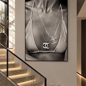 Könnte beinhalten: Ein schwarz-weiß gerahmter Wandkunstdruck, der eine Nahaufnahme einer Person in einem Bikini-Oberteil und einer langen Halskette mit einem markanten, ineinandergreifenden CC-Logo-Anhänger zeigt. Auf der Haut ist eine Bräunungslinie als CC-Logo zu sehen.