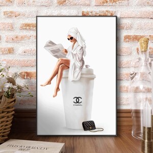 May include: A framed art print depicts a woman in a white robe and towel reading a newspaper, seated on a large white cup with the Chanel logo. A black quilted handbag with a gold chain sits nearby. The background is white.