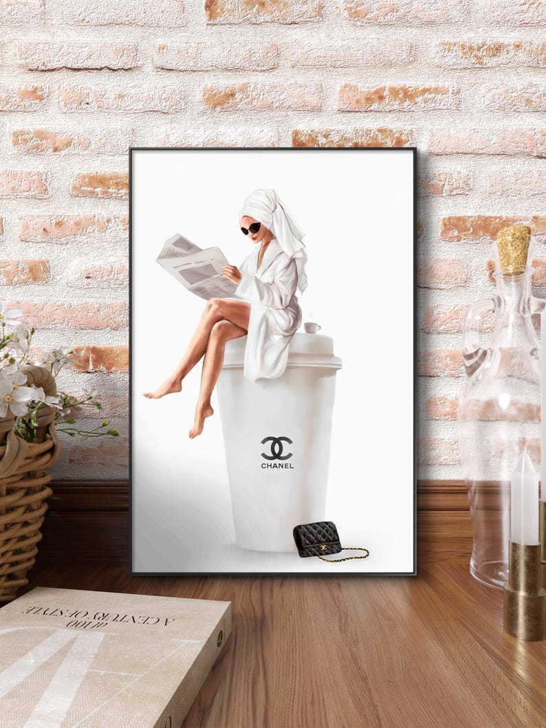 May include: A framed art print depicts a woman in a white robe and towel reading a newspaper, seated on a large white cup with the Chanel logo. A black quilted handbag with a gold chain sits nearby. The background is white.