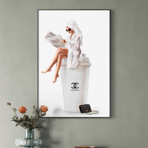 May include: A framed print depicts a woman in a white robe and towel, reading a newspaper while seated on a large coffee cup. The cup features the Chanel logo, and a black purse rests nearby. The artwork is displayed on a gray wall.