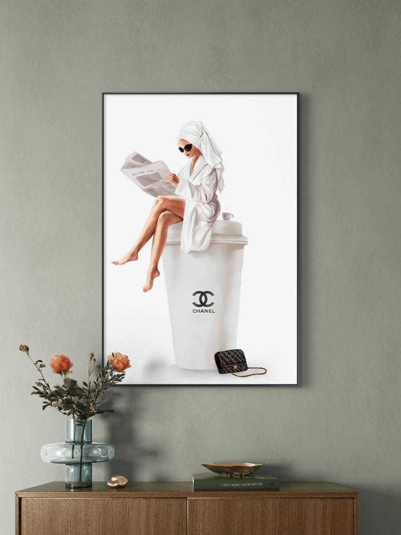 May include: A framed print depicts a woman in a white robe and towel, reading a newspaper while seated on a large coffee cup. The cup features the Chanel logo, and a black purse rests nearby. The artwork is displayed on a gray wall.