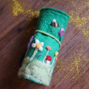 Puede incluir: Estuche enrollado de fieltro verde decorado con flores de fieltro, una abeja y un cierre de cordón verde. El estuche presenta una escena de jardín caprichosa con flores blancas, amarillas, rojas y moradas. Fondo de madera.