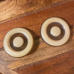 May include: Two round, cream-colored ceramic knobs with a brown concentric circle design. The knobs have a raised, circular center.