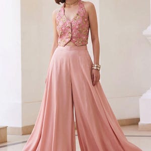 Puede incluir: Conjunto formal de dos piezas en tono rosa suave. El top, de estilo halter, presenta intrincados bordados florales y detalles de botones. El conjunto incluye pantalones palazzo de pernera ancha. Adecuado para ocasiones especiales.