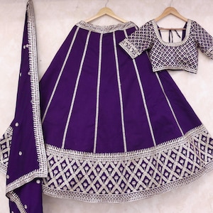 May include: A three-piece purple outfit, including a skirt, top, and scarf, hangs on wooden hangers. The skirt and top are adorned with intricate silver embroidery. The skirt features vertical lines and a geometric pattern at the hem. The top has short sleeves.