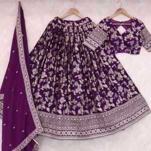 May include: A three-piece purple Indian outfit featuring a skirt, blouse, and scarf. The skirt and blouse are adorned with a floral pattern in silver. The scarf is a solid purple with silver embroidered trim. The outfit is displayed on wooden hangers.