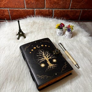 May include: A black leather-bound journal featuring a gold-colored embossed tree of life design, moon phases, and stars on the cover. The journal includes a metal latch closure and is accompanied by a metallic pen, a small Eiffel Tower model, and two miniature pots with colorful flowers.