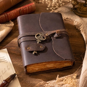 May include: A dark brown leather journal featuring a wrap-around strap closure secured with a decorative antique brass-toned metal key charm. The journal has thick, deckled-edge pages that appear aged and textured, providing a rustic appearance.