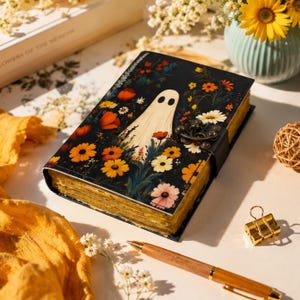 Puede incluir: Un diario rectangular con cubierta negra que presenta una ilustración central de un fantasma blanco rodeado de flores coloridas naranjas, amarillas y rojas. El libro tiene un cierre de pestillo de metal oscuro y bordes de papel envejecidos.