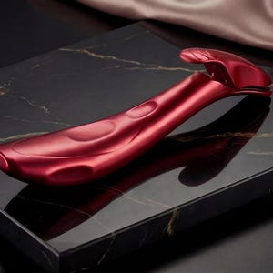 Peut inclure: Un appareil de beauté ergonomique rouge avec une poignée incurvée et une tête arrondie. L'appareil est fait d'un matériau métallique brillant et repose sur une surface en marbre noir avec des veines dorées. Un tissu rose doux est drapé en arrière-plan.