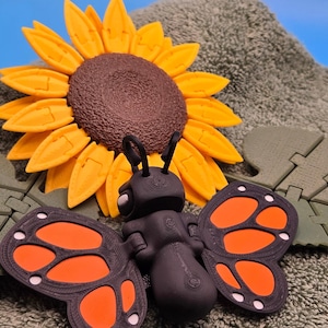 May include: A whimsical arrangement featuring a black butterfly with orange and white wing details, a yellow sunflower, and green leaf accents. The objects are set against a textured, gray background, creating a playful, nature-inspired scene.