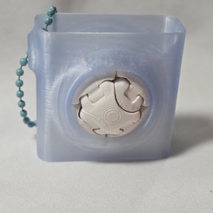 May include: A translucent blue square container with a white, gear-shaped design on the front. A teal beaded chain is attached to the side. The container is likely 3D-printed and has a smooth, slightly textured surface.