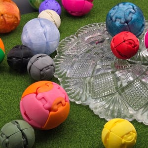 May include: A collection of multi-colored, spherical, articulated puzzle toys in various sizes and finishes, including solid, translucent, and metallic colors, scattered alongside a clear, decorative, pressed glass dish with a diamond pattern.