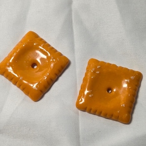 Peut inclure: Deux crackers carrés orange avec une finition brillante sont présentés sur une surface blanche. Chaque cracker a un trou central et un bord strié. Les crackers sont d'une couleur orange vif.