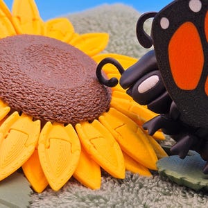 May include: A whimsical, handcrafted diorama featuring a large, yellow and brown sunflower and a black butterfly with orange markings. The butterfly appears to be interacting with the sunflower, creating a charming, nature-themed scene.