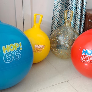 May include: Four colorful hopper balls. A blue ball with "HOP! 66" text, a yellow ball with "HOP! 44", a clear ball with glitter, and a red ball with "HOP! 55". Each ball has a handle.