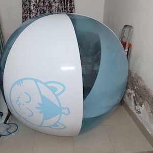 Puede incluir: Una gran bola inflable esférica tipo zorb con paneles alternos de color azul claro translúcido y blanco opaco. El panel blanco muestra una ilustración gráfica en azul claro de una cara de gato estilizada con los ojos cerrados y orejas pequeñas.