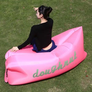 May include: A bright pink inflatable lounger with the word "doughnut" in green script. The lounger is on a grassy surface. The lounger is designed for outdoor relaxation.