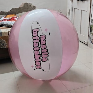 May include: A large, round, inflatable beach ball featuring alternating panels of translucent pink and opaque white. The white panel displays the text 'nastila inflatables' in a bold, dark font, encircled by a thin black ring with small star accents.