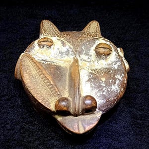 Op de afbeelding: Een gesneden houten masker met een gestileerd dierengezicht. Het masker is voornamelijk bruin met delen van witte en beige verf. Het heeft grote, ronde ogen, een lange neus en een gedetailleerde mond. Het masker heeft twee kleine, puntige oren aan de bovenkant.