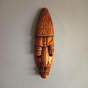 Op de afbeelding: Een gesneden houten masker met een bruine afwerking. Het masker heeft een lange, ovale vorm met gedetailleerde gelaatstrekken, waaronder een prominente neus, ogen en mond. De bovenkant van het masker heeft een hoedachtig ontwerp met kruisarcering.