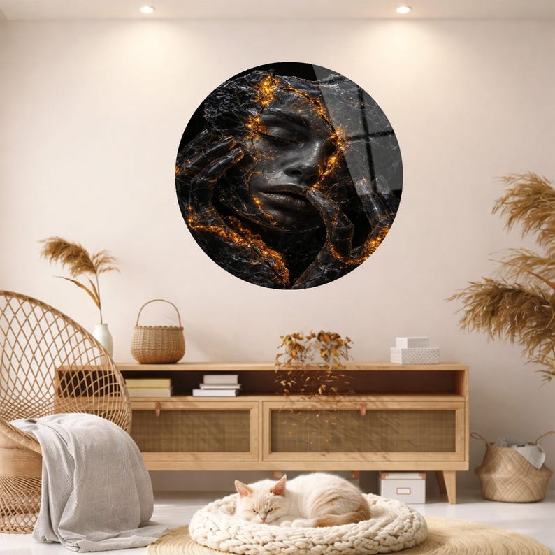 May include: A round wall art piece featuring a black and gold abstract face. The artwork has a textured appearance with gold accents. The image is displayed on a light-colored wall, above a wooden cabinet. A cat is sleeping in a round bed.