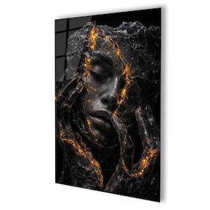 May include: A black and gold abstract art print featuring a woman's face. The face is partially obscured by a cracked, textured surface with glowing gold accents. The woman's hands are near her face, adding to the artistic composition.
