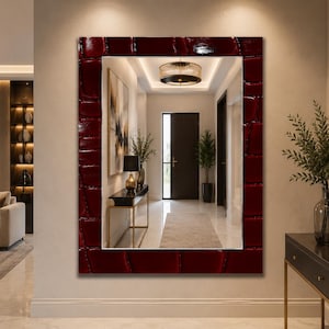 May include: A rectangular wall mirror featuring a wide, deep red frame with a glossy, textured crocodile skin pattern. The mirror surface reflects a bright, modern hallway with a console table, a lamp, a potted plant, and a dark wooden door.