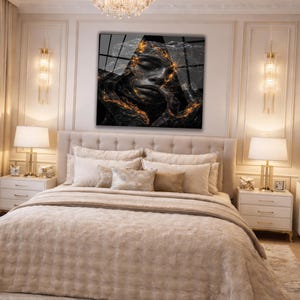 May include: A bedroom scene featuring a large bed with a beige tufted headboard and matching bedding. Above the bed hangs a black and white artwork with gold accents. The room is decorated in neutral tones with gold accents, including lamps and wall sconces.