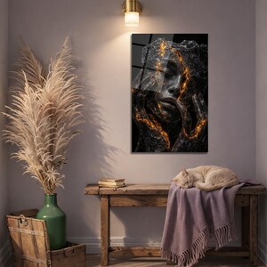 May include: A framed art print featuring a woman's face with a cracked, textured appearance. The cracks are highlighted with gold, creating a striking contrast against the dark background. The artwork is displayed on a wall.
