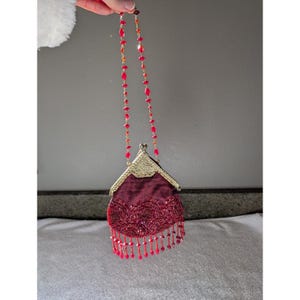 May include: A burgundy evening bag with a gold clasp and beaded strap. The bag features intricate beadwork and a fringe of red beads. The strap is made of red and orange beads. The bag is held up by a hand.