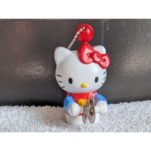 May include: A white Hello Kitty figurine with a red bow, red ball, and silver chain. The figurine is wearing a blue and red outfit and holding two silver cymbals. The figurine is on a white surface.