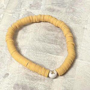 May include: A light yellow beaded bracelet with a white bead accent. The bracelet is made of small, flat, disc-shaped beads strung together. The white accent bead has a gold-colored letter "K" on it.
