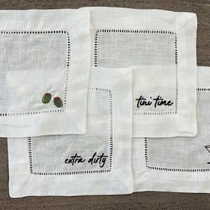 Op de afbeelding: Een set van vier vierkante witte linnen cocktailservetten met ajourzoom. De servetten zijn geborduurd met zwarte tekst "tini time" en "extra dirty", samen met kleine illustraties van groene olijven en een martiniglas.