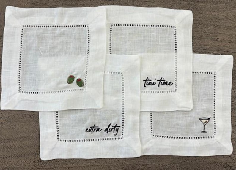 Op de afbeelding: Een set van vier vierkante witte linnen cocktailservetten met ajourzoom. De servetten zijn geborduurd met zwarte tekst "tini time" en "extra dirty", samen met kleine illustraties van groene olijven en een martiniglas.