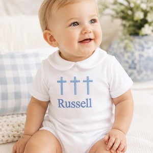 May include: A white short-sleeved romper with a Peter Pan collar, featuring an embroidered design of three light blue crosses above the name Russell in matching blue thread. The garment has a simple, clean aesthetic with a classic round neckline.