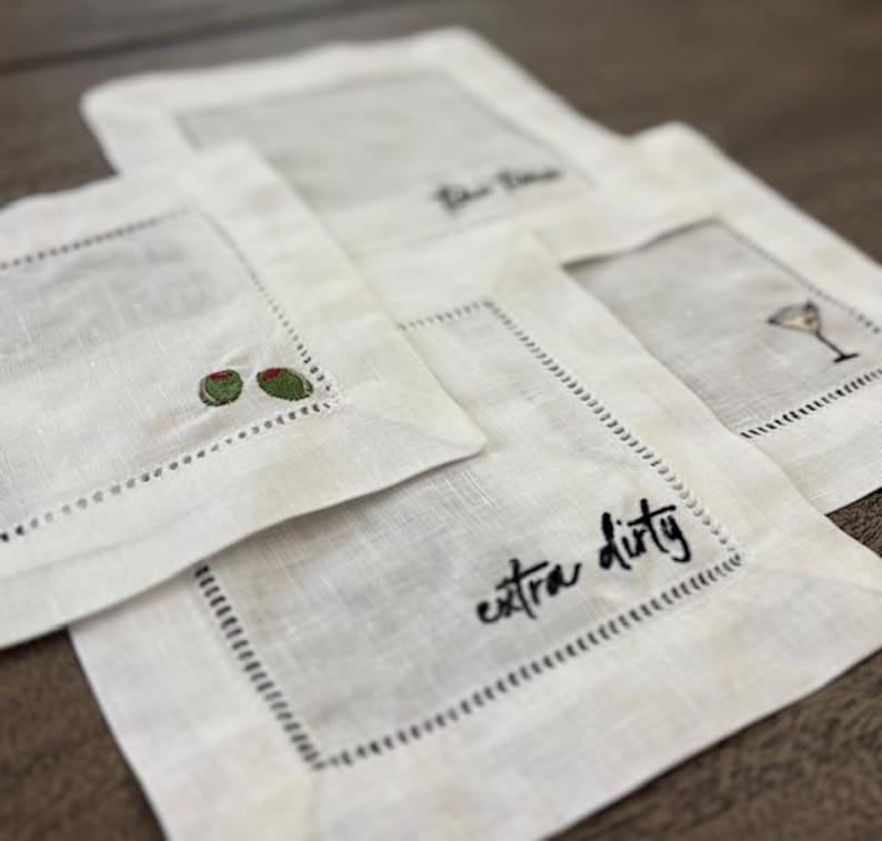 Op de afbeelding: Een set van vier vierkante witte linnen cocktailservetten met ajourzoom. Elke servet heeft uniek borduurwerk, waaronder twee groene olijven, een martiniglas en de in zwart geborduurde tekst die extra dirty luidt.