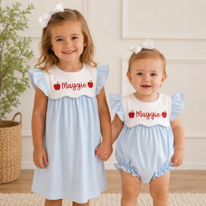 Pode incluir: Um vestido azul claro e um macacão a combinar, ambos com um colarinho branco recortado bordado com o nome Maggie em letra vermelha, ladeado por dois pequenos ícones de maçãs vermelhas e laços brancos para o cabelo.